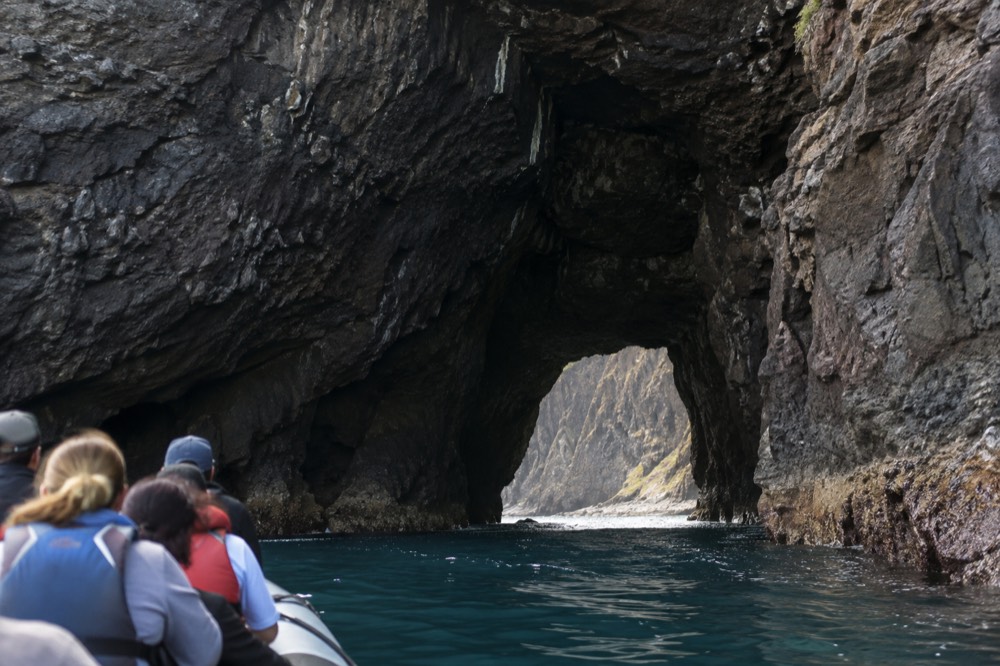 Raft passengers in life vests approaching a wide sea cave entrance on a calm morning