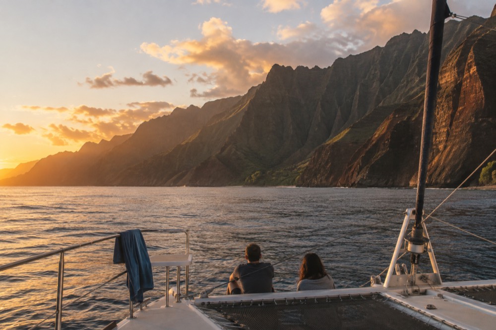 A catamaran under way on calm water near a tropical coastline at sunset.