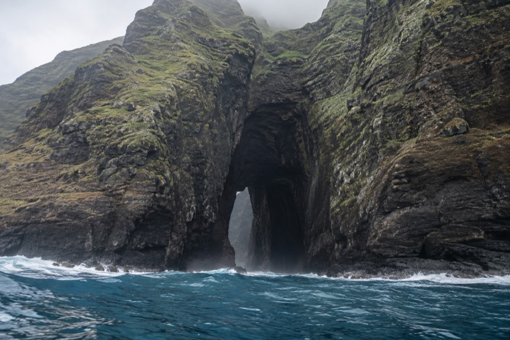 A tall sea cave opening in a sheer green cliff face along a remote Pacific coastline