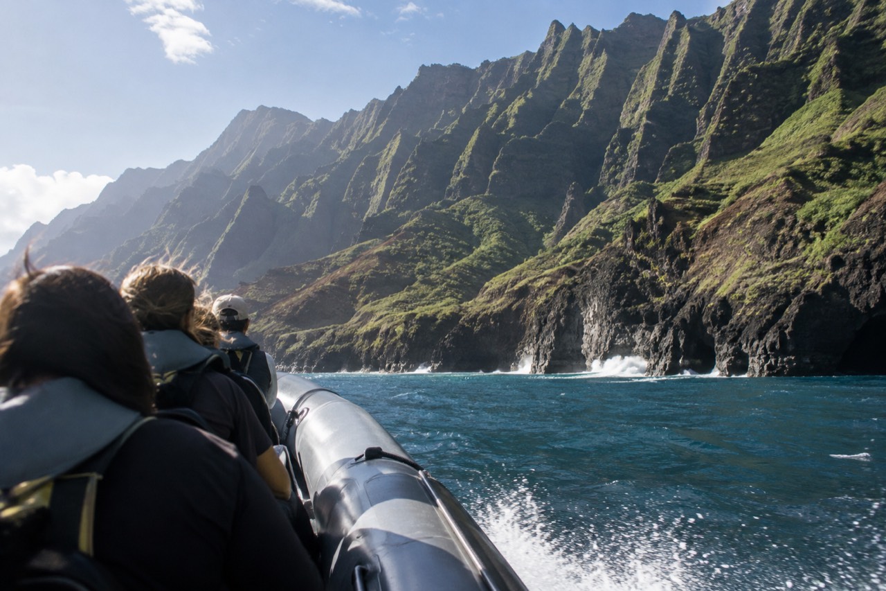 A rigid hull inflatable raft cruising close to the tall green sea cliffs of the Napali Coast under a bright sky