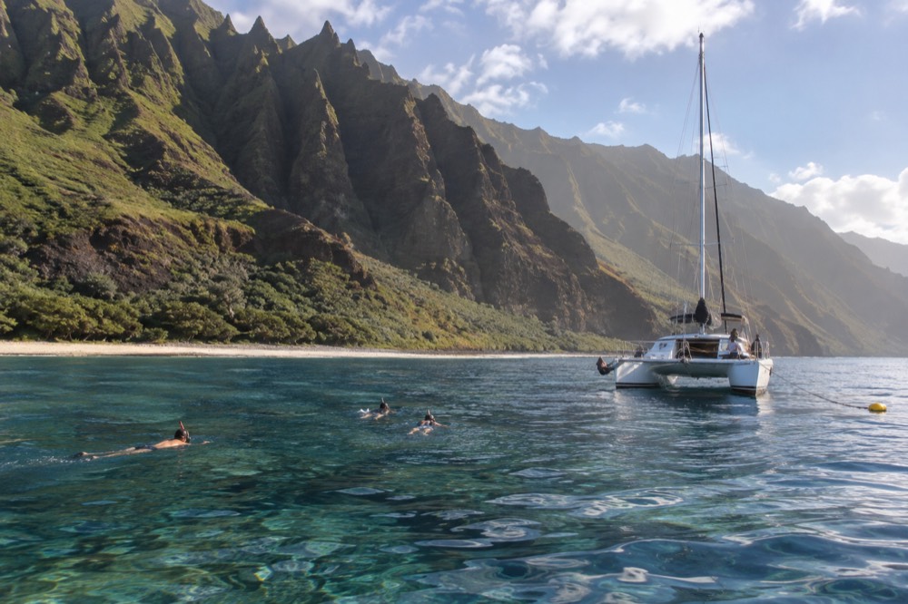 A catamaran anchored near a reef with snorkelers in clear water below sea cliffs