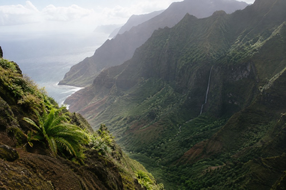 Sea cliffs and a waterfall along a green Hawaiian coastline seen from a distance