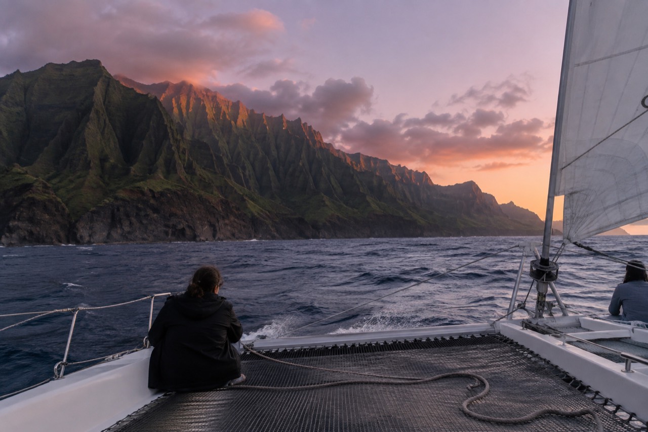 A catamaran under sail along a steep green sea cliff coastline at dusk