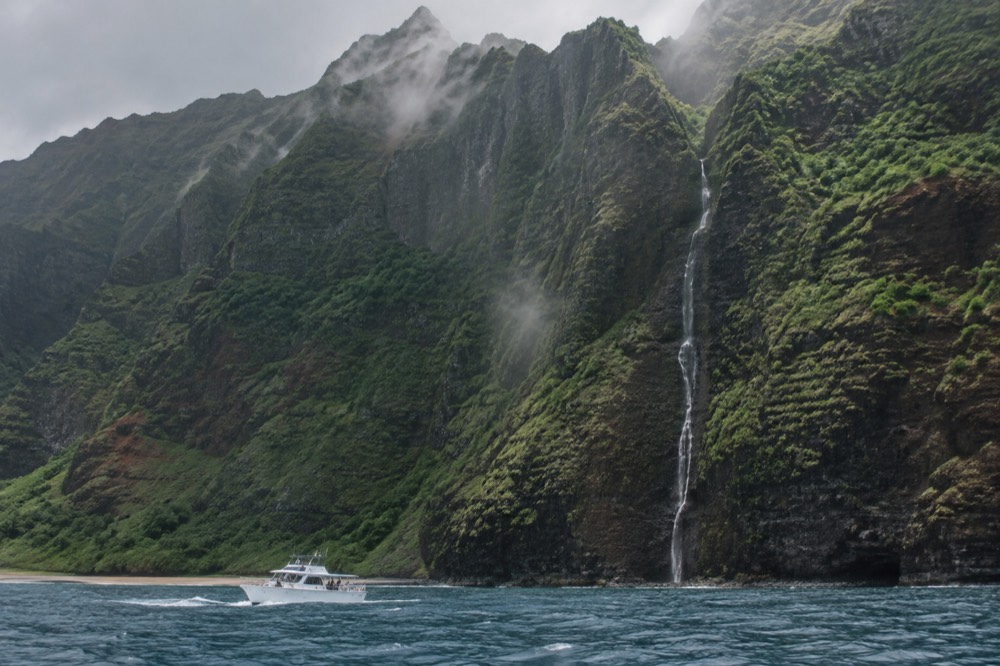 A small boat cruising near tall green sea cliffs along the Napali Coast with a waterfall visible on the rock face