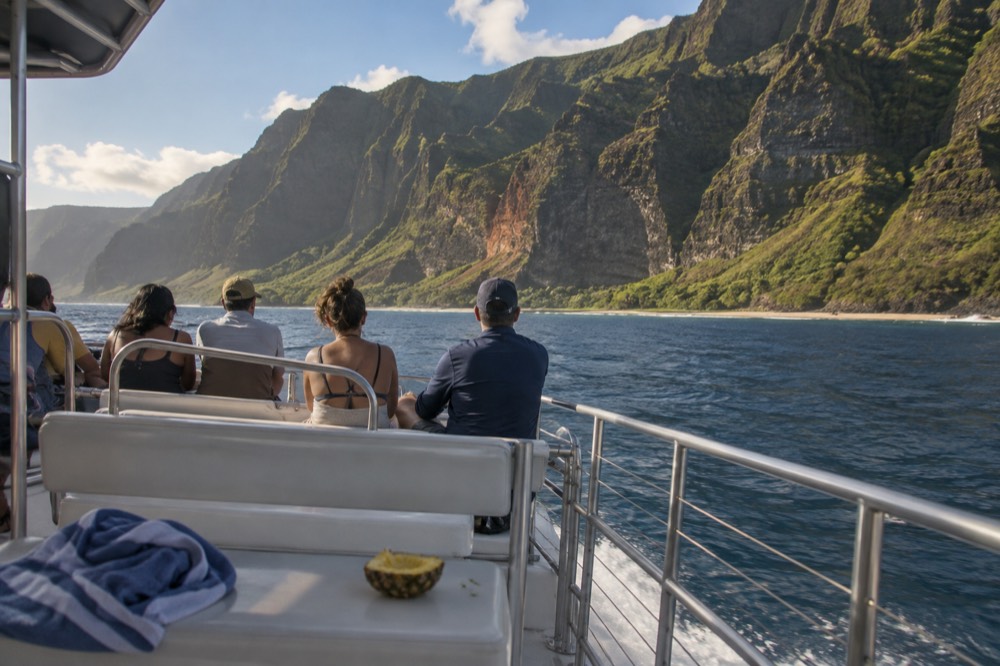 A power catamaran cruising past tall green sea cliffs with passengers seated on the forward deck