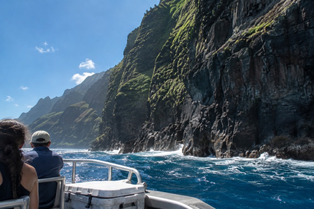 A small boat cruising close to tall vegetated sea cliffs at midday