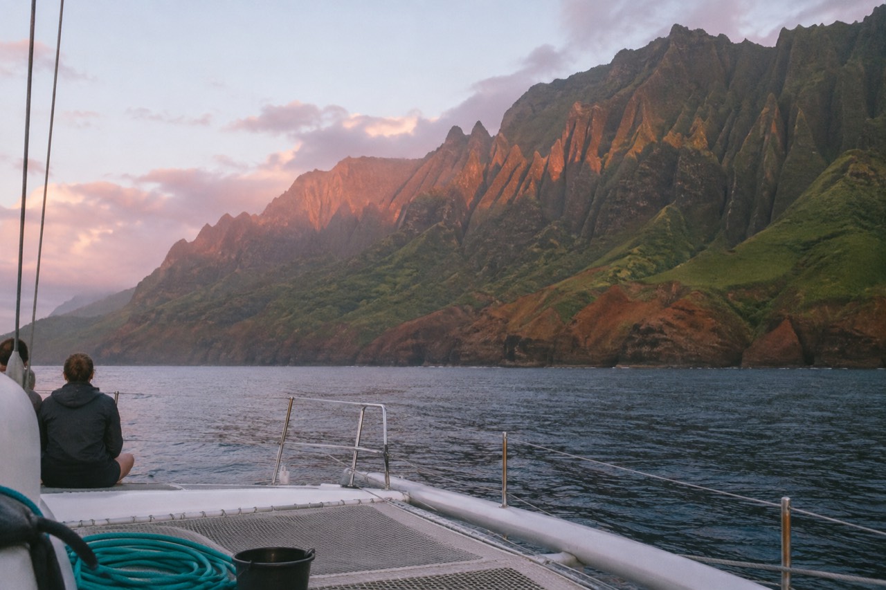 A catamaran cruising along the green sea cliffs of the Napali Coast on a calm morning