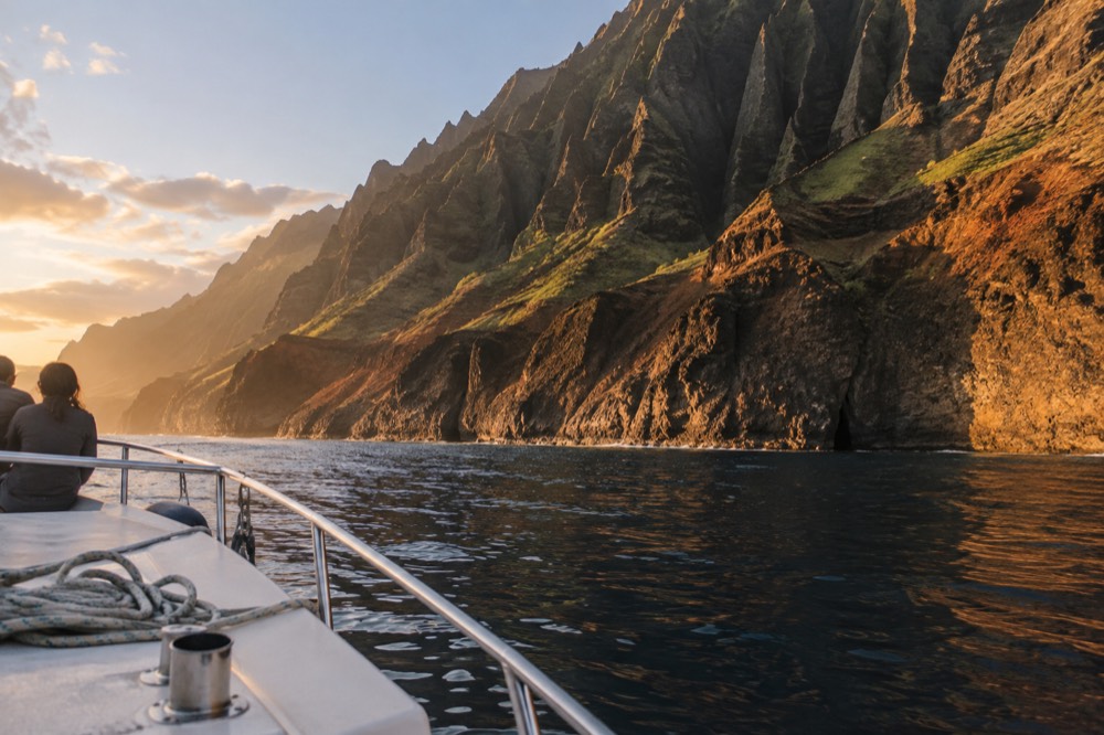 A catamaran under sail near dark cliff walls at golden evening light