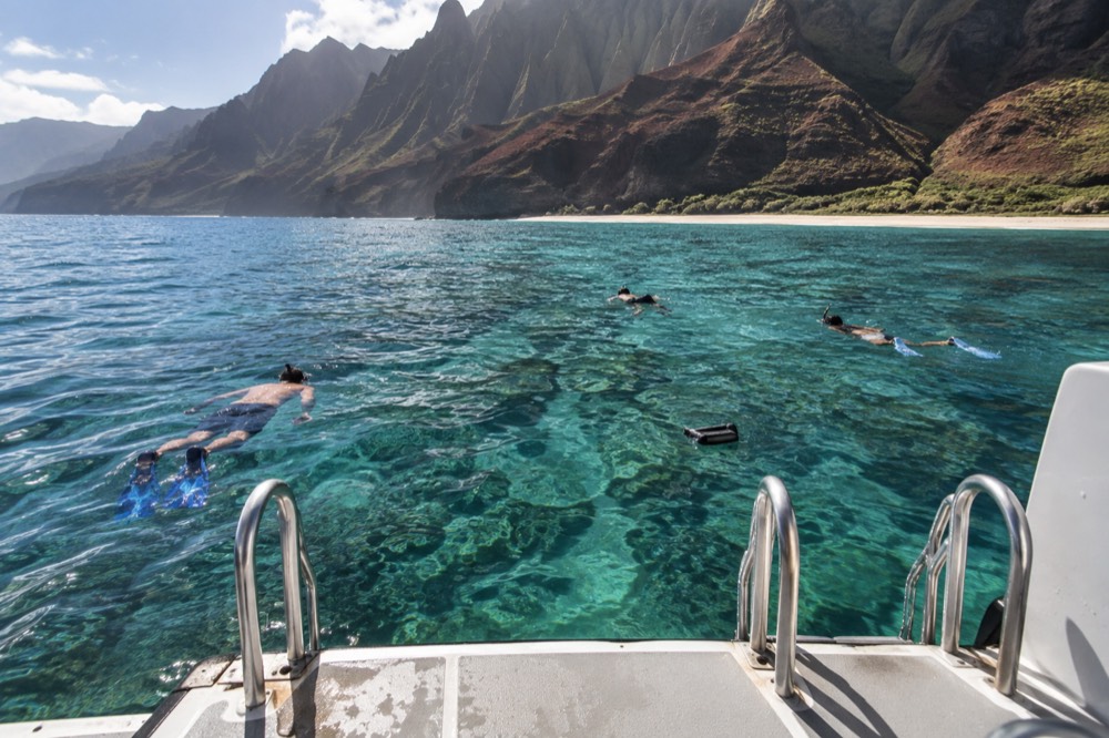 A power catamaran anchored near tall cliffs while passengers snorkel in clear water