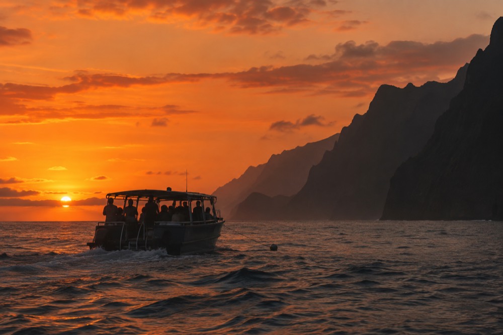 A boat silhouetted against an orange sunset sky near coastal cliffs