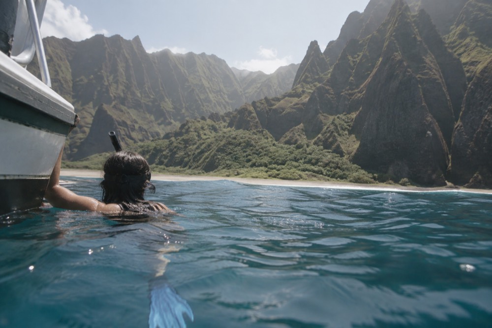 A snorkeler at the surface near a boat in calm blue water beside a green coastline