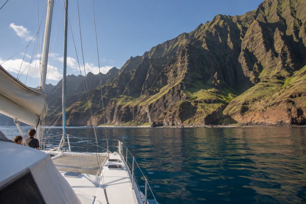 A sailing catamaran passing below tall green sea cliffs on a calm morning