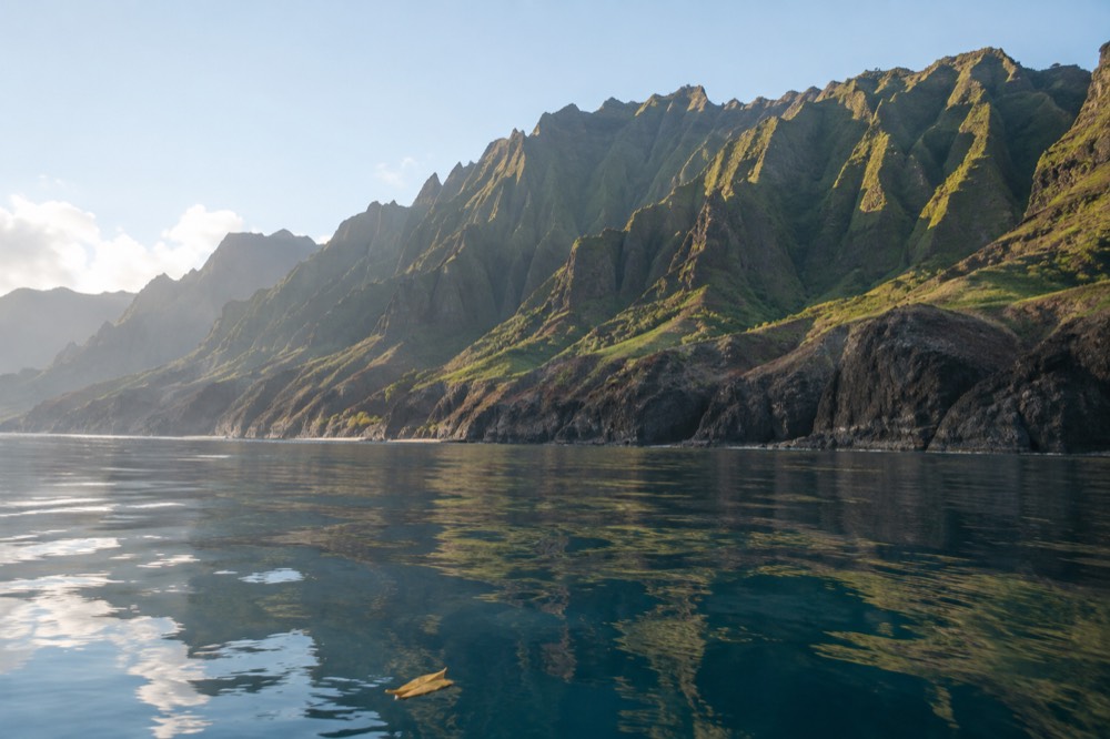 Glassy calm water along the Napali Coast in morning light, cliffs reflected on the surface.