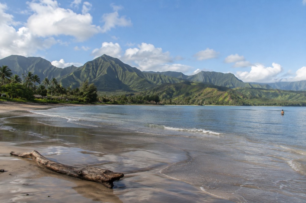 A wide north-facing bay with a long sand beach and steep green ridges under scattered clouds.