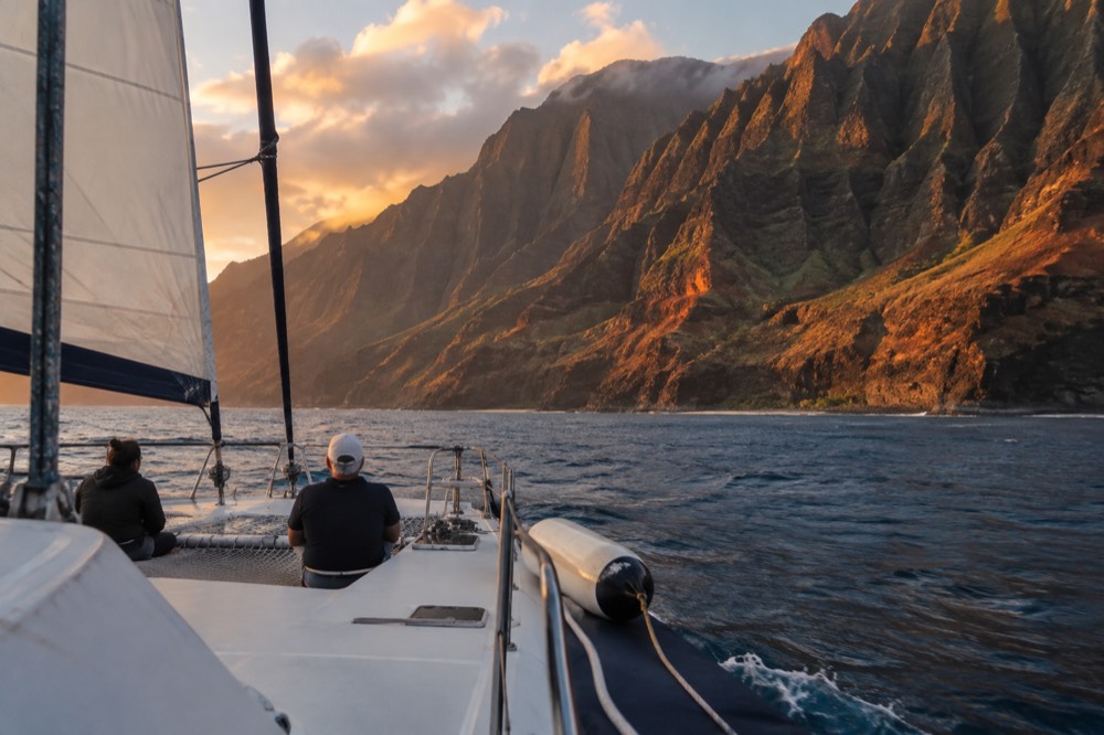 A small sailing catamaran under working canvas on open water off the Napali Coast cliffs