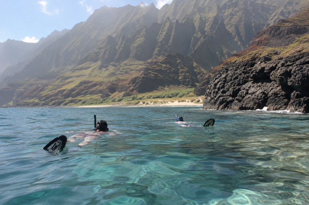 Snorkellers in clear water above a reef near dark volcanic rocks