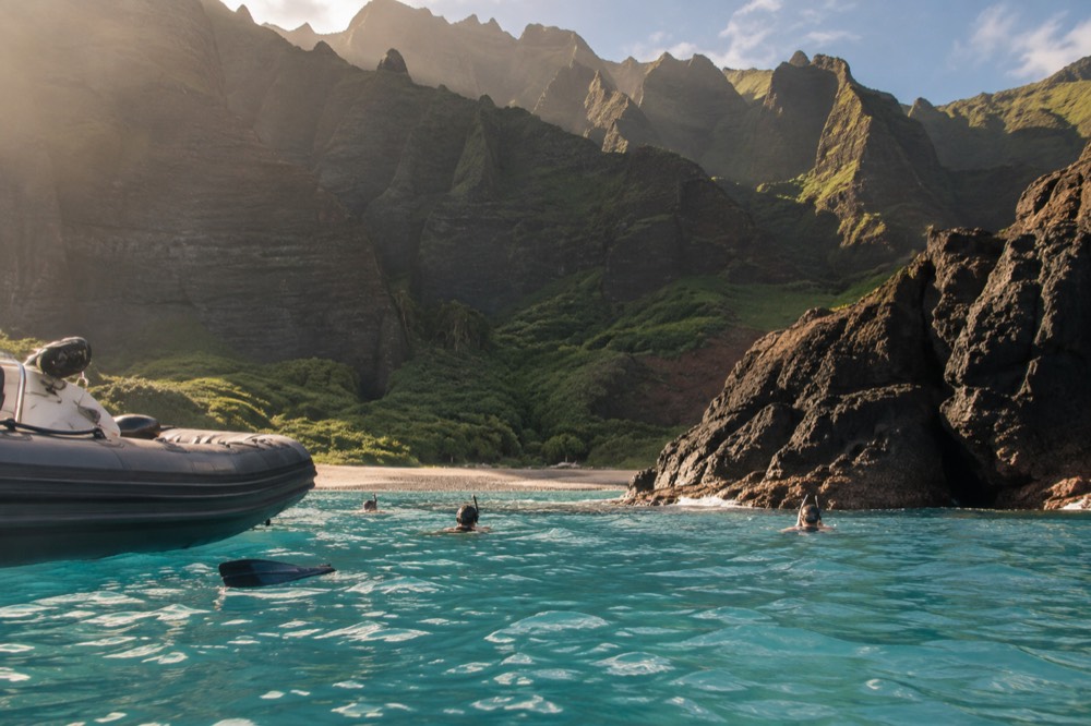 A small rigid inflatable boat at anchor in clear water beside a rocky shoreline with snorkelers nearby
