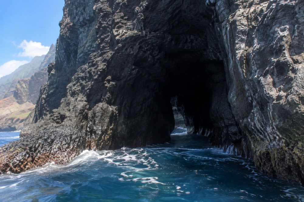 A cave opening in a cliff face at water level with blue ocean water flowing at its mouth