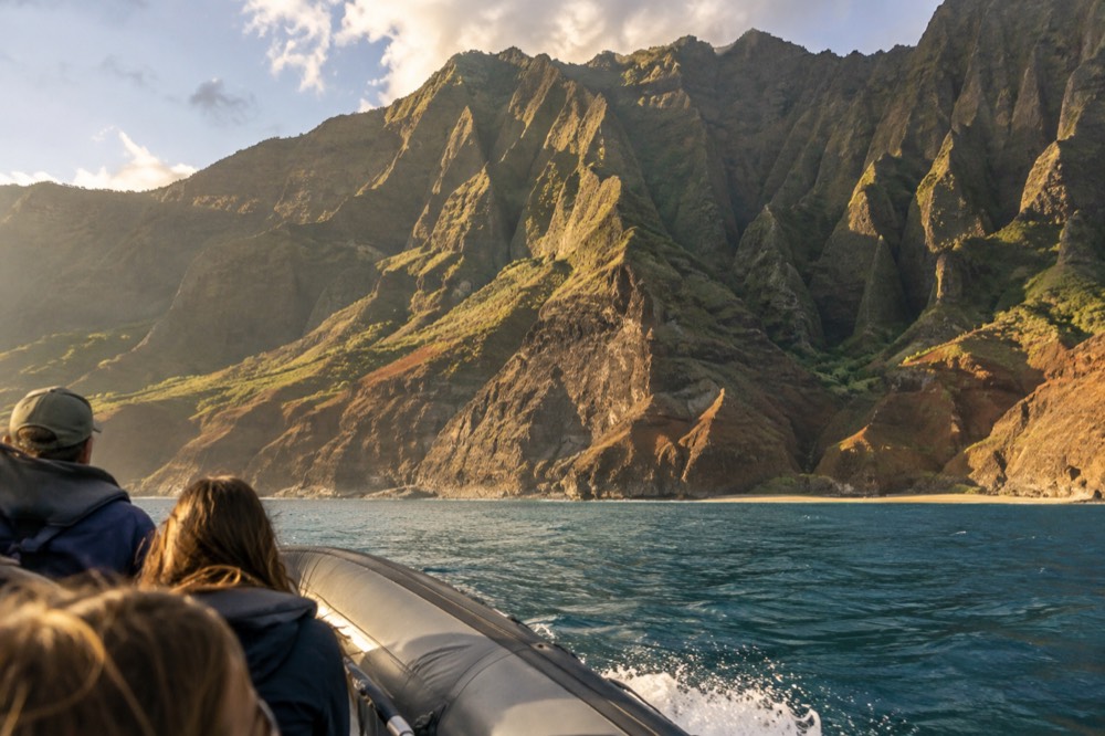 A rigid inflatable boat cruising along a stretch of tall eroded sea cliffs in golden afternoon light