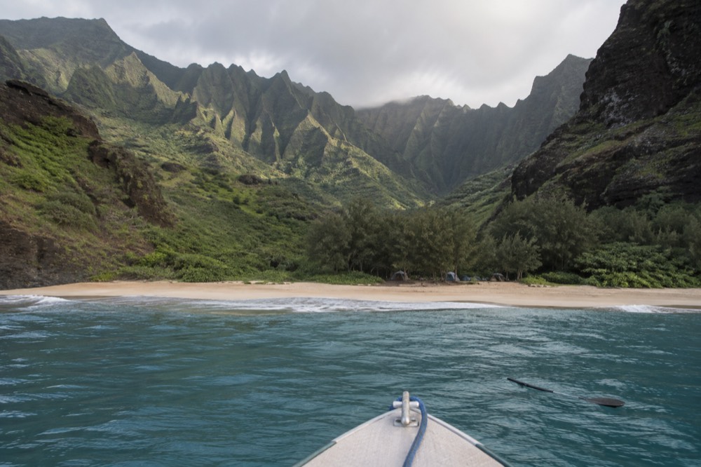 Close on a whole-boat charter's bow pointing toward the narrow sand crescent of Kalalau Beach below green valley